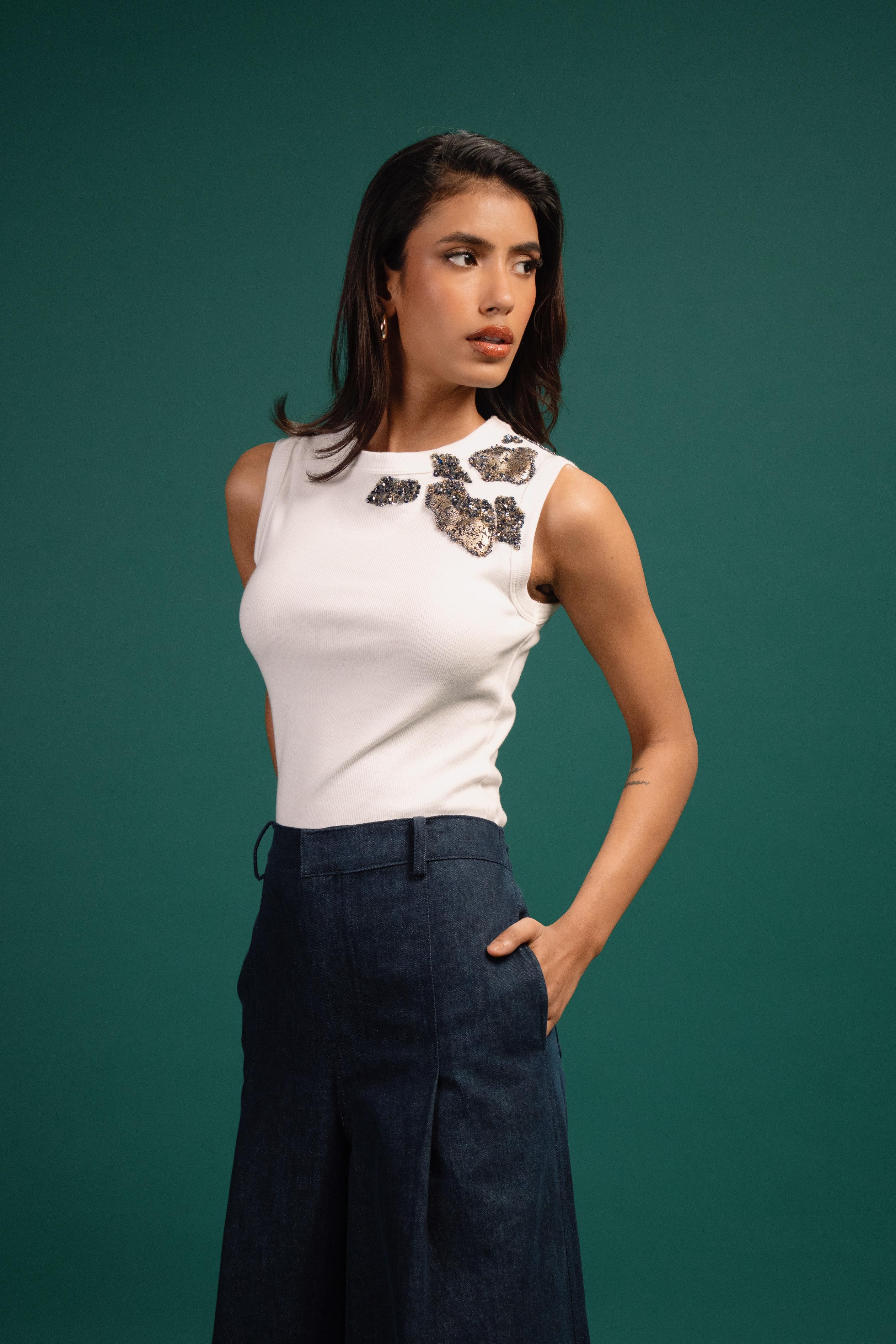Woman wearing a white sleeveless top with decorative elements and dark pants against a green background